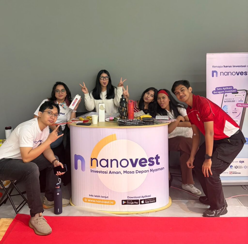 Group of six young people posing at a Nanovest promotional booth with a curved counter and banner on the right.