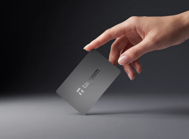 r - Blog Nanovest Close-up of a hand placing a black Tangem card onto a flat gray surface, logo visible on the card.